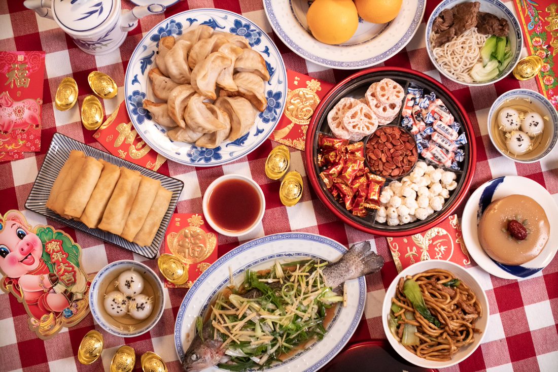 8 lucky Lunar New Year foods including dumplings, spring rolls, oranges, whole fish, year cake and sweet rice dumplings.